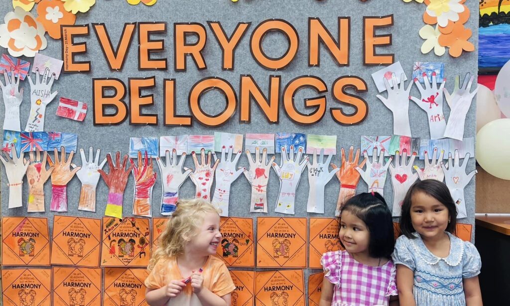 Three small children standing in front of a classroom artwork that says "Everyone Belongs"